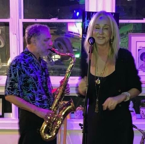 David Kreimer playing tenor sax next to guest vocalist Barbara Steel at a microphone, under purple stage light.